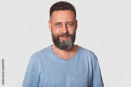 Relaxed calm middle-aged man standing isolated over light grey background looking at camera. Pleasant handsome smiling model has dark beard, wearing casual light blue t shirt. Emotions concept. © sementsova321