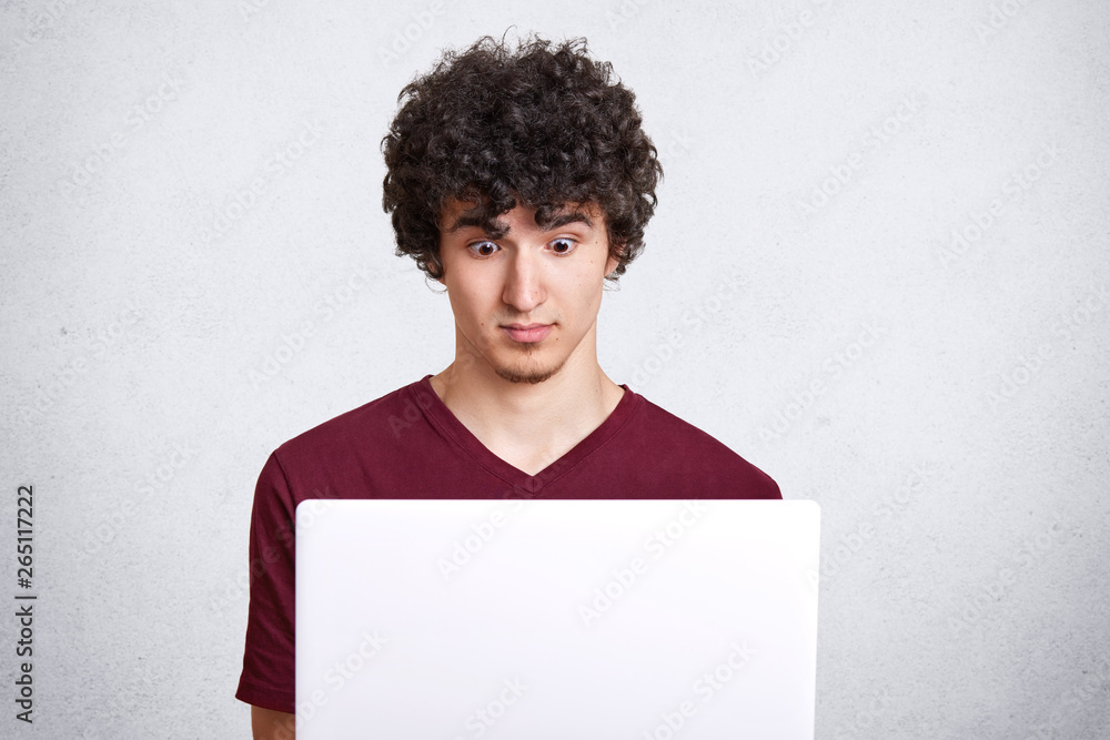 Photo of handsome male with dark curly hair, freelancer works from home, sits in front of laptop with astonished facial expression and wide opened eyes, wears casual maroon t shirt, isolated on white. Photo of handsome male with dark curly hair, freelancer works from home, sits in front of laptop with astonished facial expression and wide opened eyes, wears casual maroon t shirt, isolated on white.