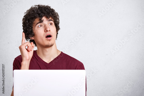 Close up portrait of young handsome freelancer man working with laptop computer at home, pointing finger up, think about new successful idea, sitting against white studio wall with opened mouth. photo