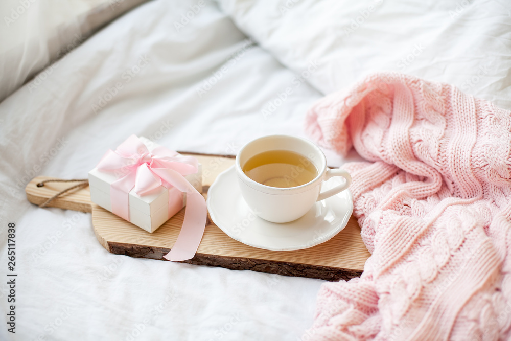 White mug with tea, gift box with ribbon on the bed. Breakfast in bed. Cozy. Pink plaid. Cotton. White mug with tea, gift box with ribbon on the bed. Breakfast in bed. Cozy. Pink plaid. Cotton.