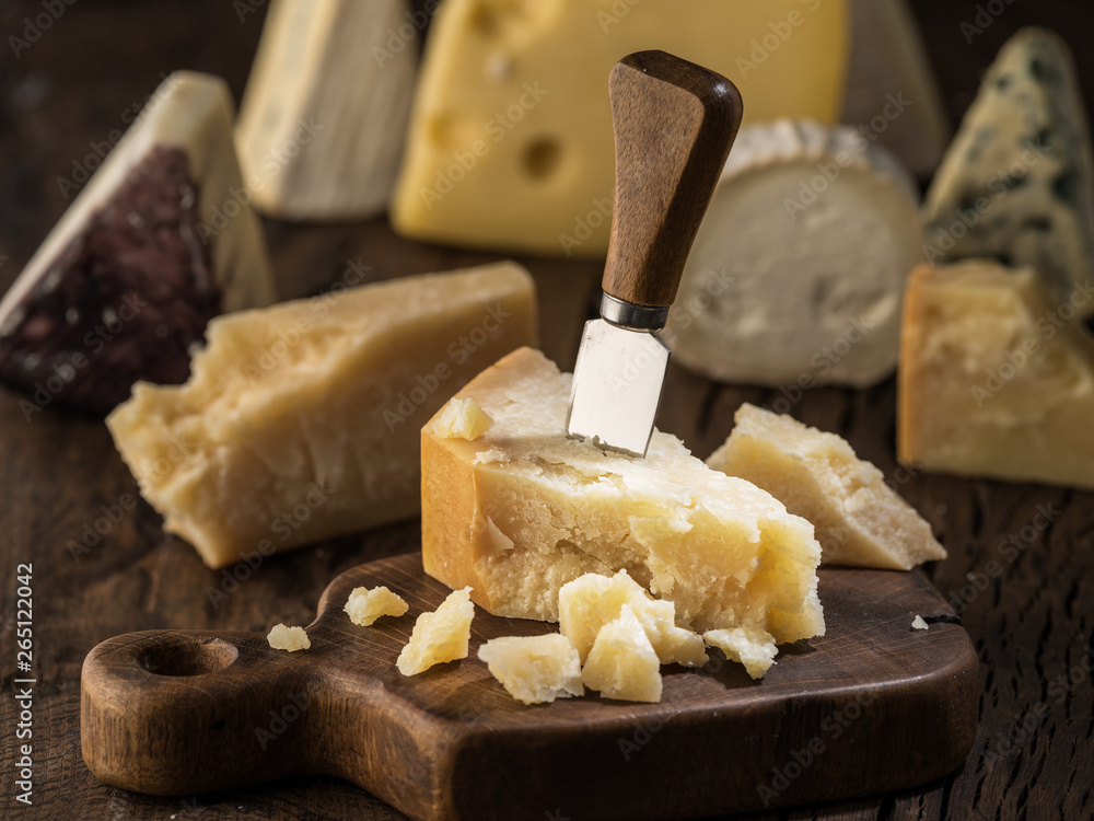 Piece of Parmesan cheese on the wooden board. Assortment of different cheeses at the background. Piece of Parmesan cheese on the wooden board. Assortment of different cheeses at the background.