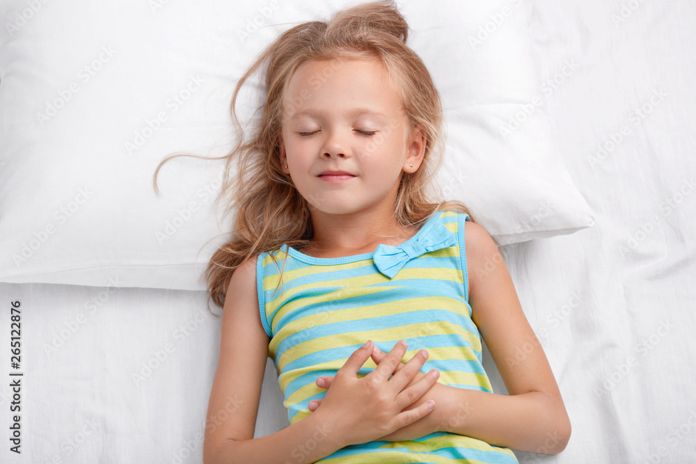 Charming lovely small child with messy hair, healthy skin, keeps hands together, lies on comfortable bed, has pleasant dreams during afternoon nap, Children, good night and sleeping concept. Charming lovely small child with messy hair, healthy skin, keeps hands together, lies on comfortable bed, has pleasant dreams during afternoon nap, Children, good night and sleeping concept.