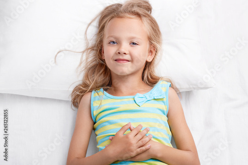 Pretty small child has pleasant look, charming smile, light hair, blue eyes, healthy skin, keeps hands on stomach, dressed in striped pyjamas, looks directly at camera, rests in bedroom alone.