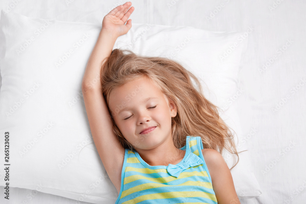 Good looking small female child with pleasant smile, stretches in bed, keeps eyes shut, dressed in casual nightclothes, closes eyes, sees pleasant dreams during afternoon rest, poses in white bed Good looking small female child with pleasant smile, stretches in bed, keeps eyes shut, dressed in casual nightclothes, closes eyes, sees pleasant dreams during afternoon rest, poses in white bed
