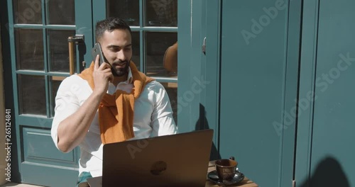 Bearded businessman working outdoors on laptop while talking smartphone, sunny day in outdoors cafe photo