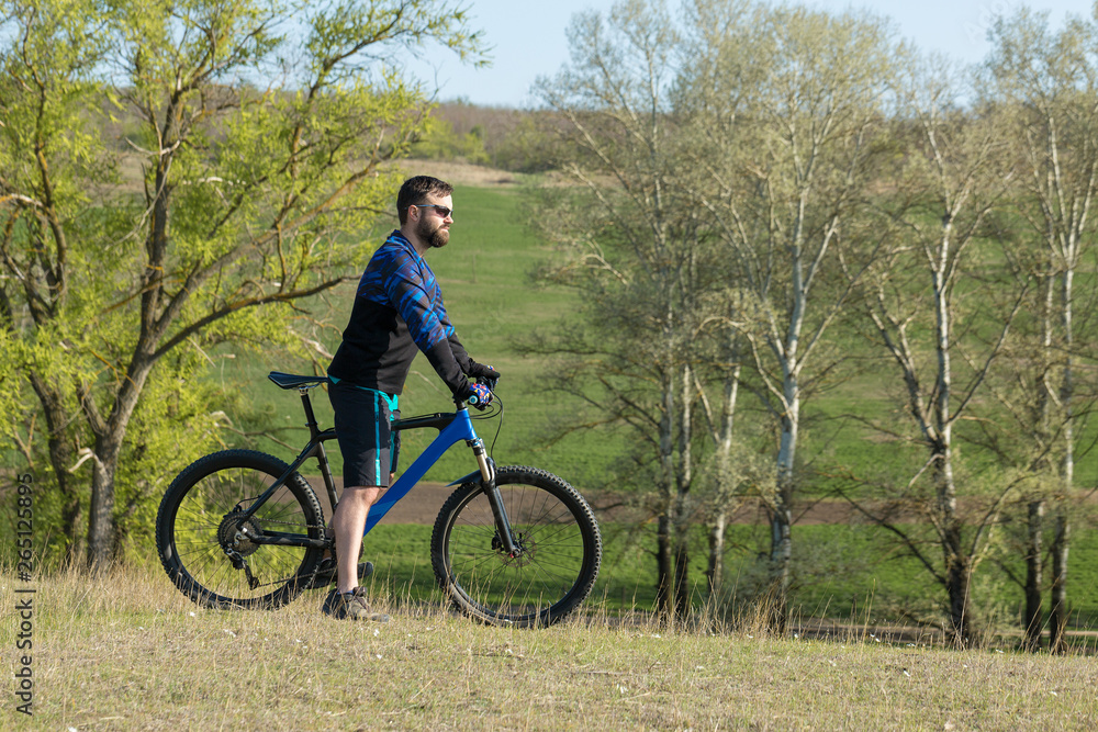Cyclist in shorts and jersey on a modern carbon hardtail bike with an air suspension fork rides off-road on green hills near the forest Cyclist in shorts and jersey on a modern carbon hardtail bike with an air suspension fork rides off-road on green hills near the forest