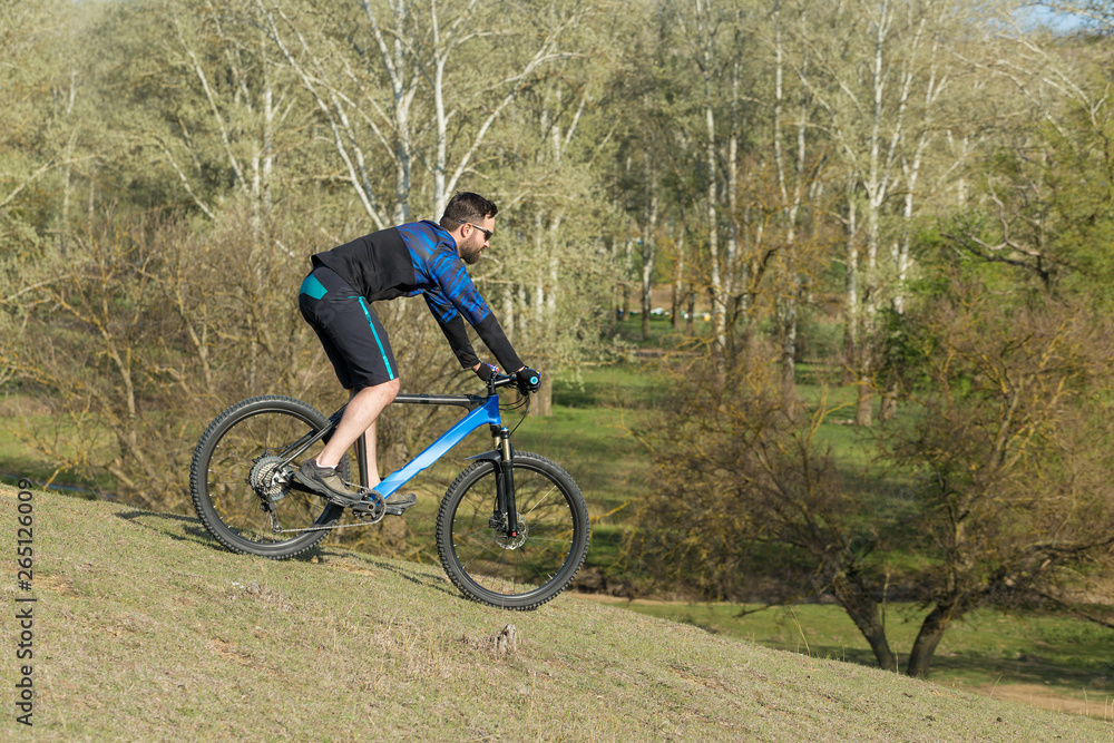 Cyclist in shorts and jersey on a modern carbon hardtail bike with an air suspension fork rides off-road on green hills near the forest