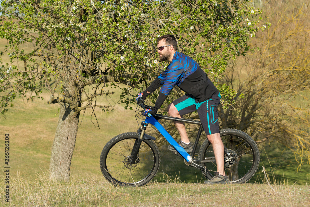 Cyclist in shorts and jersey on a modern carbon hardtail bike with an air suspension fork rides off-road on green hills near the forest Cyclist in shorts and jersey on a modern carbon hardtail bike with an air suspension fork rides off-road on green hills near the forest