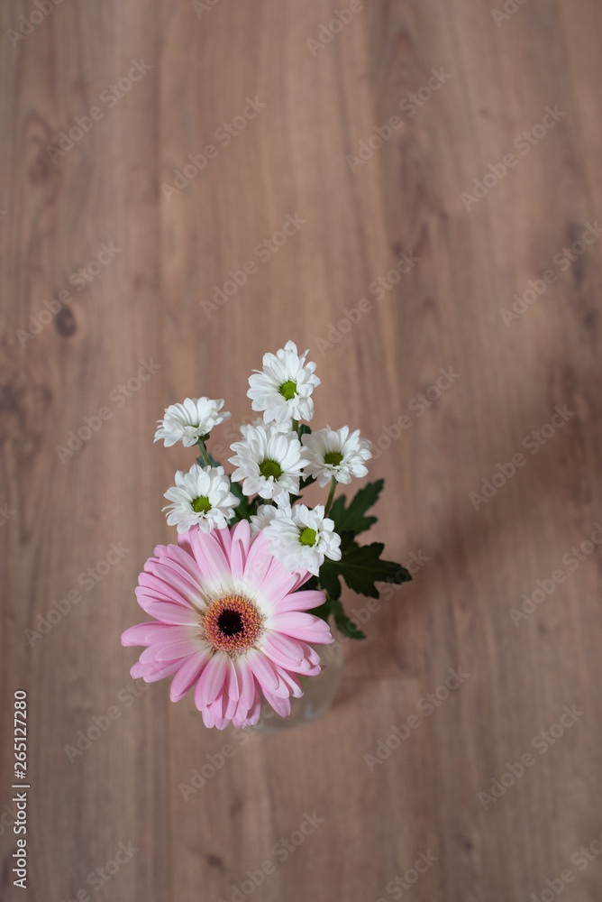 Fototapeta premium Blumenstrauss aus einer rosa pinken Gerbera und weissen Blumen in Glasvase auf Holzboden unten mittig platziert von oben fotografiert im Hochformat