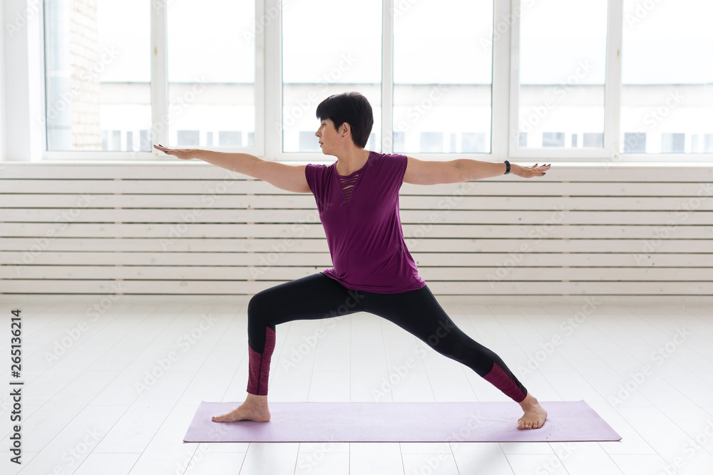 People, yoga, sport and healthcare concept - Middle-aged woman standing in Warrior over white background