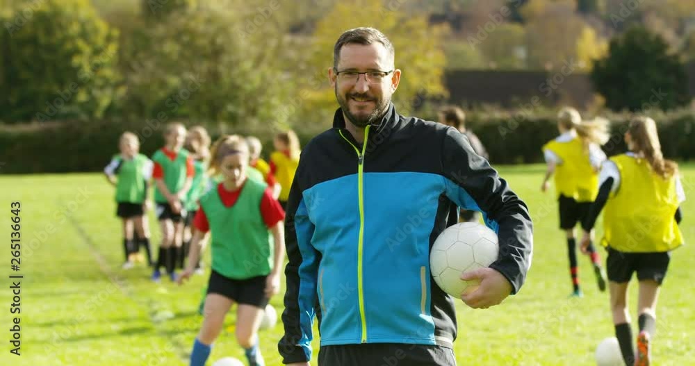 4K Portrait smiling sports coach holding a football, with girls' soccer team training in background 4K Portrait smiling sports coach holding a football, with girls' soccer team training in background