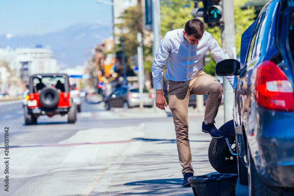A strong businessman is bending his knee while trying to change a flattie on his car with his lug wrench. A strong businessman is bending his knee while trying to change a flattie on his car with his lug wrench.