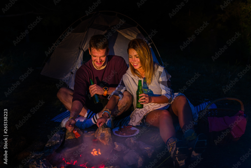 Fototapeta premium Young couple baking sausages on the campfire and drinking beer in the forest hill in the dusk
