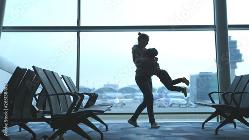Playful mother raises her son and holds him in arms looking through window in airport.