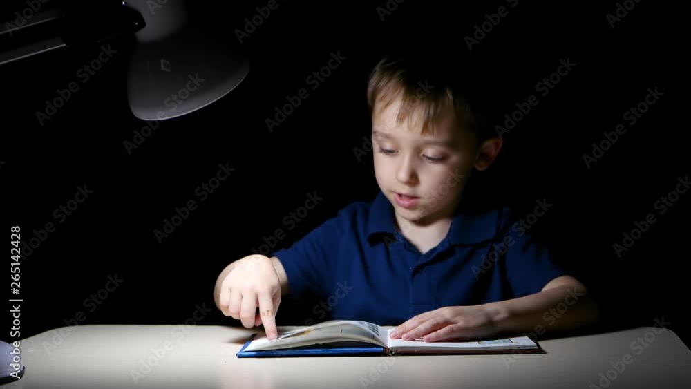 A child with his finger points to the drawings in the book at home, before going to sleep, he sits at a table lit by a lamp A child with his finger points to the drawings in the book at home, before going to sleep, he sits at a table lit by a lamp