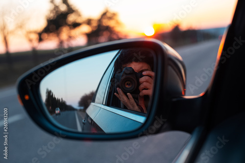 Young photographer taking selfie picture in a mirror of car with professional dslr camera, on background of sunset. © Lalandrew