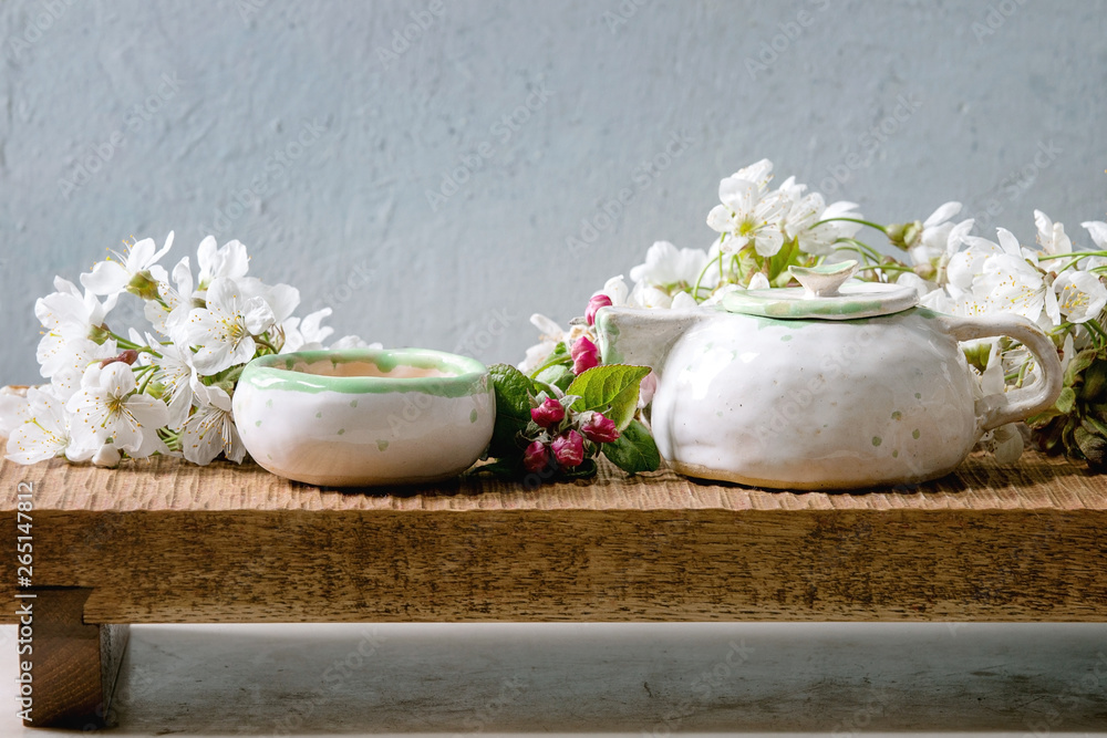 Craft handmade ceramic teapot and cup with hot tea decorated by spring blossom cherry branches on japanese style wooden serving tray on white marble table. Craft handmade ceramic teapot and cup with hot tea decorated by spring blossom cherry branches on japanese style wooden serving tray on white marble table.