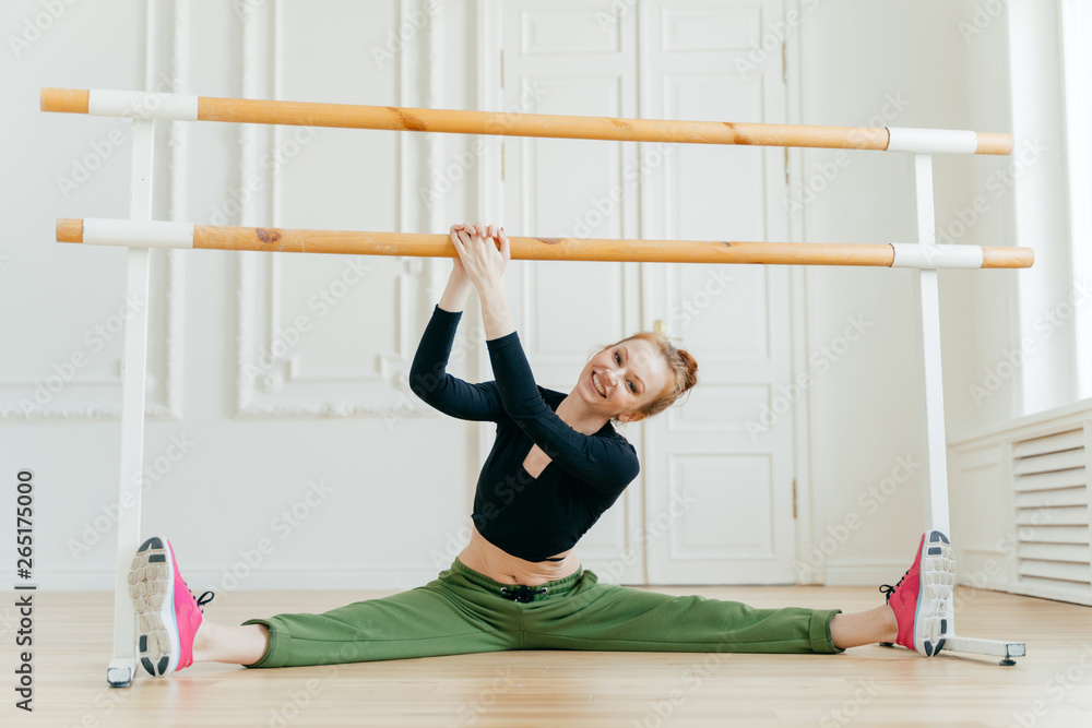 Naklejka premium Caucasian female ballerina spreads legs sideways, shows splits, holds hands on barres, has ballet class, wears sportswear and sneakers, has perfect figure, tilts head on right, stretches herself.