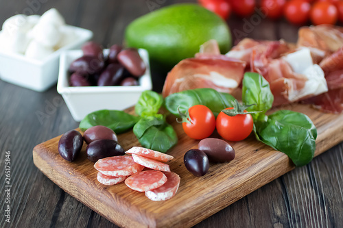 Prosciutto, bread, olives, walnut, mozzarella, salami, basil and cherry tomatoes on brown wooden board. Mediterranean kitchen.