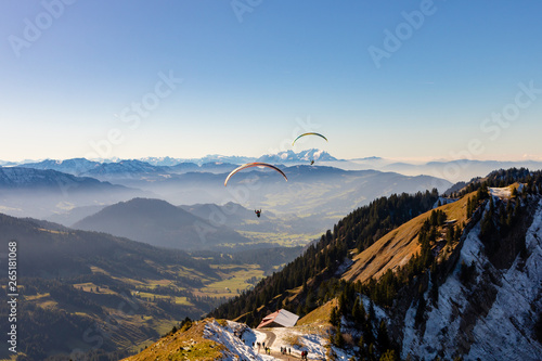 Paragliders flying straight to a valley over a hut with people around in Oberstaufen, Germany.