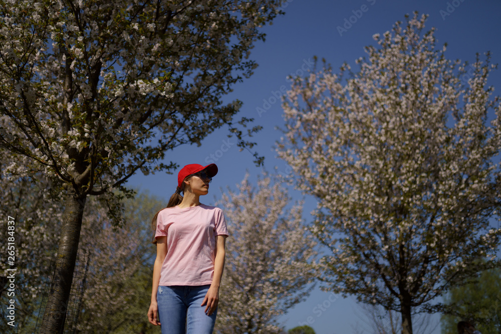 Successful business woman enjoys her leisure free time in a park with blossoming sakura cherry trees wearing jeans, pink t-shirt and a fashion red cap Successful business woman enjoys her leisure free time in a park with blossoming sakura cherry trees wearing jeans, pink t-shirt and a fashion red cap