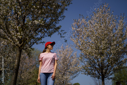 Successful business woman enjoys her leisure free time in a park with blossoming sakura cherry trees wearing jeans, pink t-shirt and a fashion red cap