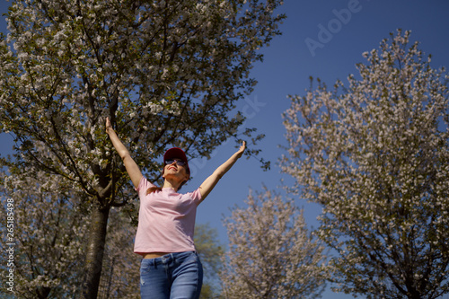 Successful business woman enjoys her leisure free time in a park with blossoming sakura cherry trees wearing jeans, pink t-shirt and a fashion red cap