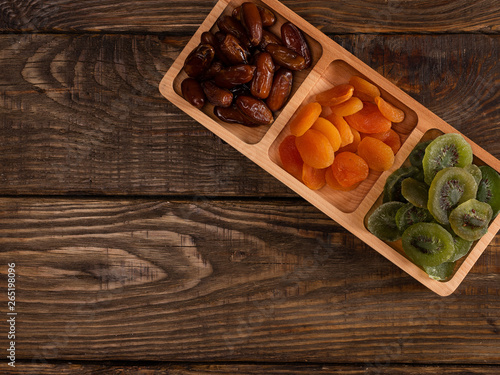 Dates, dried apricots and kiwis in a Compartmental dish on a dark wooden table.