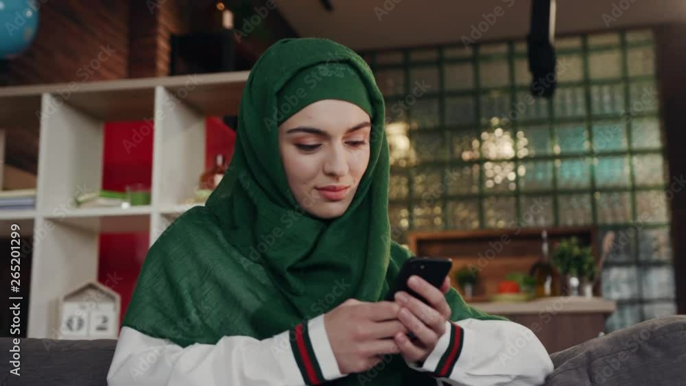 Elegant attractive female muslim in hijab smiling straight to camera, using mobile smartphone searching online information while sitting on sofa couch relaxing. Technologies concept Elegant attractive female muslim in hijab smiling straight to camera, using mobile smartphone searching online information while sitting on sofa couch relaxing. Technologies concept