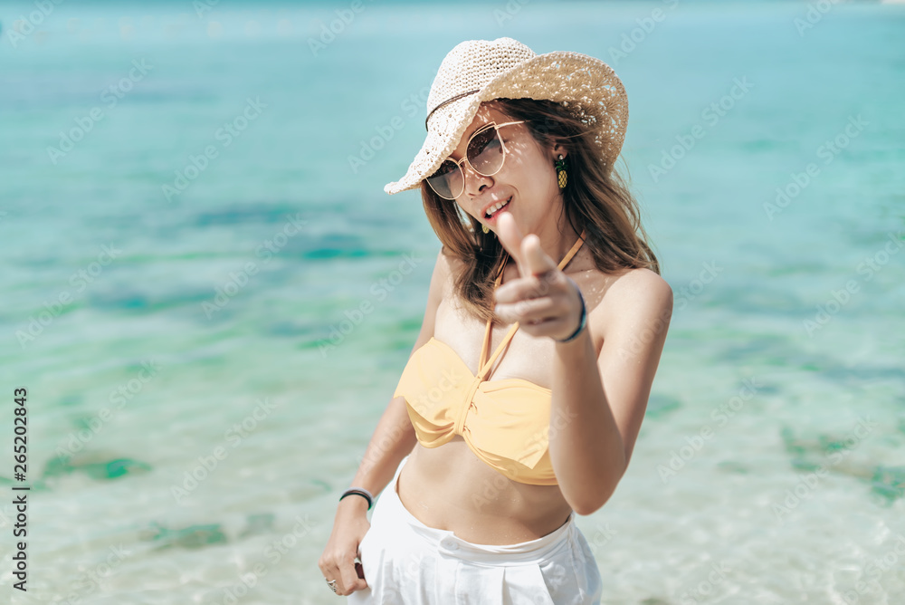Cheerful girl wears bikini and white hat posing emotionally with sea waves and horizon on background.Pretty woman in sunglasses and orange attire standing on sandy beach on blue sea background