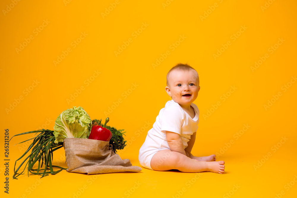 baby girl sitting with bag of vegetables on yellow isolate background, space for text. the concept of healthy eating.organic food baby girl sitting with bag of vegetables on yellow isolate background, space for text. the concept of healthy eating.organic food