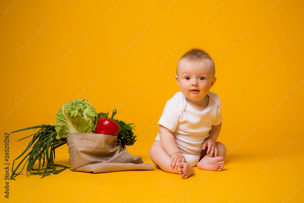 baby girl sitting with bag of vegetables on yellow isolate background, space for text. the concept of healthy eating.organic food baby girl sitting with bag of vegetables on yellow isolate background, space for text. the concept of healthy eating.organic food