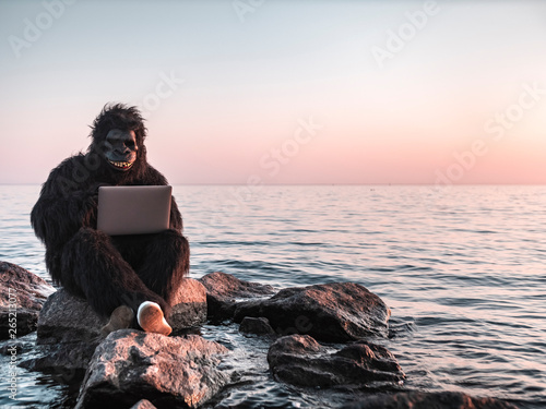 A man in a monkey costume at sunset near the sea works on a computer against the backdrop of a modern skyscraper. Animal programmer does the job