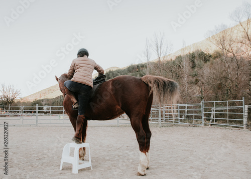 Back view of female in helmet mounting on obedient horse in enclosure on ranch