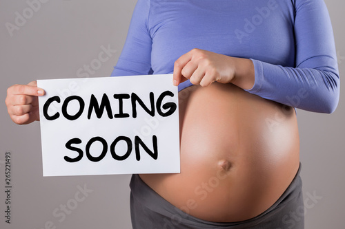 Image of close up stomach of pregnant woman holding paper with text coming soon on gray background.