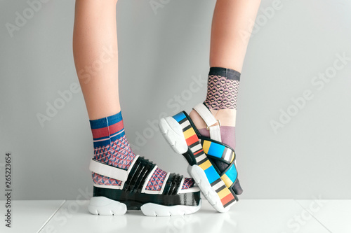 Beautiful female legs in mismatched trendy socks standing in two different fashionable high wedge leather sandals on white surface. Odd disargonized  young girl wearing high sole summer stylish shoes.