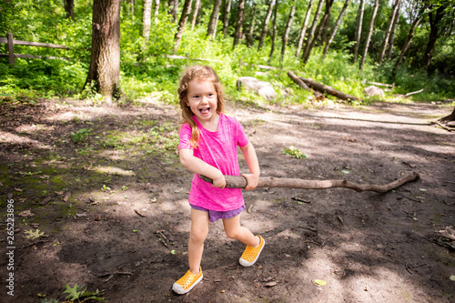 A toddler aged girl hiking in a forest and dragging a large stick.
