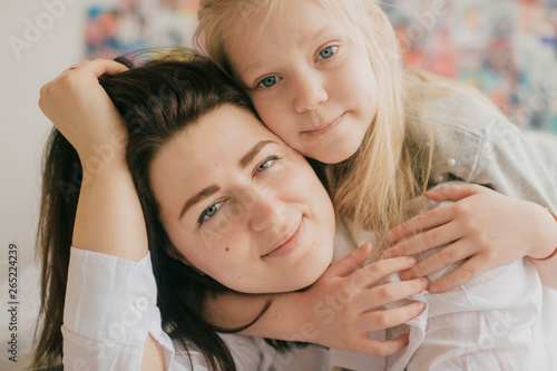 Lifestyle soft focus portrait of happy mom hugs her adorable daughter on white bed. Indoor happy family portrait of smiling mother and her daughter lying together on bed with decorative wall behind.