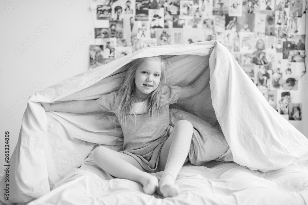 Funny little girl sitting on white bed and holding blanket above her