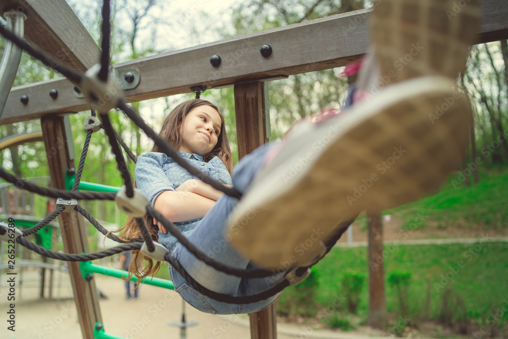 Obraz premium Cute girl having fan on playground. Charming little brunette girl in jeans suit hanging on grid and looking at camera on playground in park
