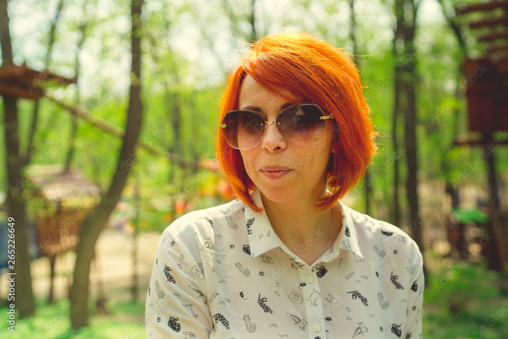 Young woman with red hair, sunglasses, among the trees. Portrait of young attractive woman with fashionable hairstyle sitting in Park Young woman with red hair, sunglasses, among the trees. Portrait of young attractive woman with fashionable hairstyle sitting in Park