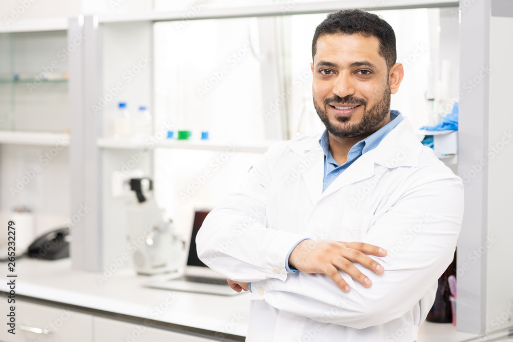 Smiling handsome young middle-eastern lab worker with beard in white coat crossing arms on chest and looking at camera in laboratory