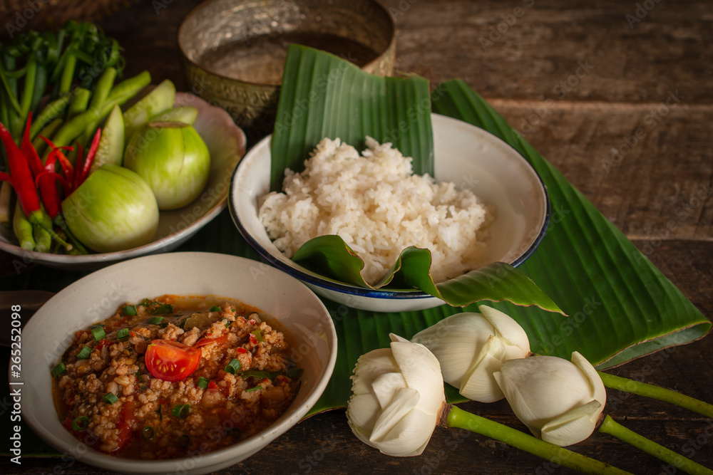 Thai Northern Style Pork and Tomato Chili Relish, nam prik ong in white bowl on wood table there are side dishes of fresh vegetables, cooked rice, water bowl and flower placed around. Thai Northern Style Pork and Tomato Chili Relish, nam prik ong in white bowl on wood table there are side dishes of fresh vegetables, cooked rice, water bowl and flower placed around.