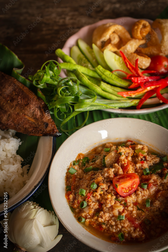 Thai Northern Style Pork and Tomato Chili Relish, nam prik ong in white bowl on wood table there are side dishes of fresh vegetables, cooked rice and flower placed around.