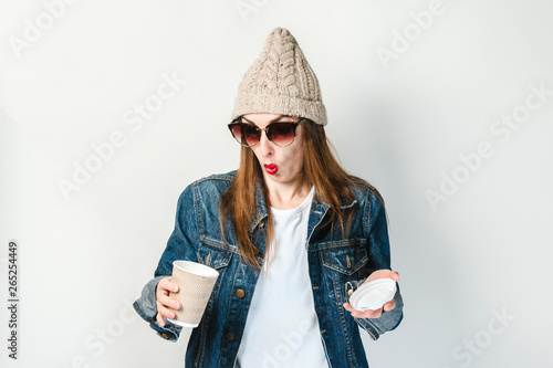 Young girl in a denim jacket, glasses and a hat looks into an empty paper cup for coffee or tea on a white background. Concept breakfast, coffee at work, coffee shop, recharging, over coffee.
