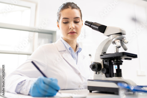 Serious thoughtful young woman in lab coat sitting at table and making records about research in laboratory