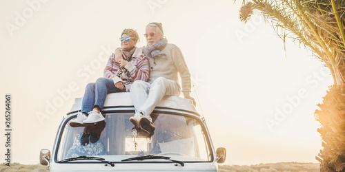Wanderlust and travel destination happiness concept with old senior beautiful couple sitting and enjoying the outdoor freedom on the roof of vintage van vehicle together - sun backlight