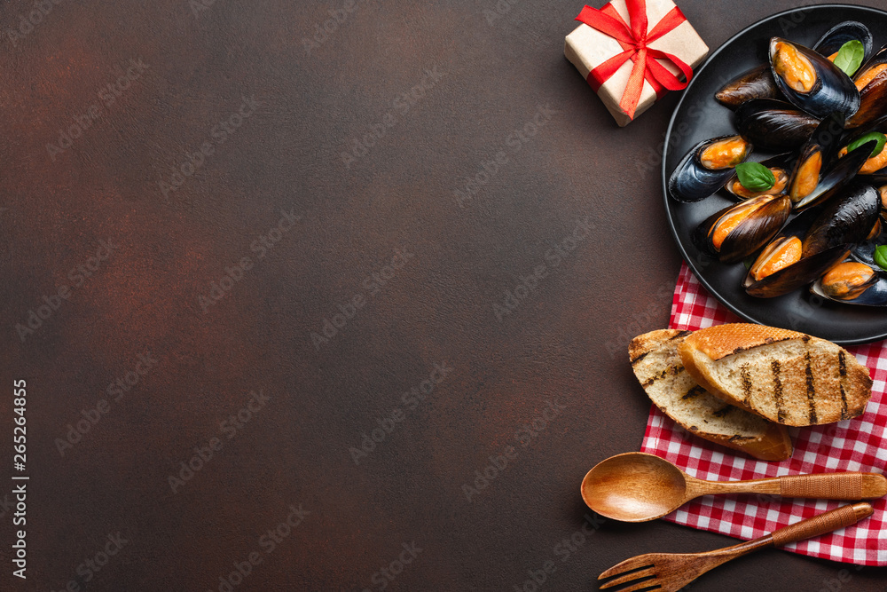 Seafood mussels and basil leaves in a black plate with towel, bread slices, wooden spoon and fork on rusty background