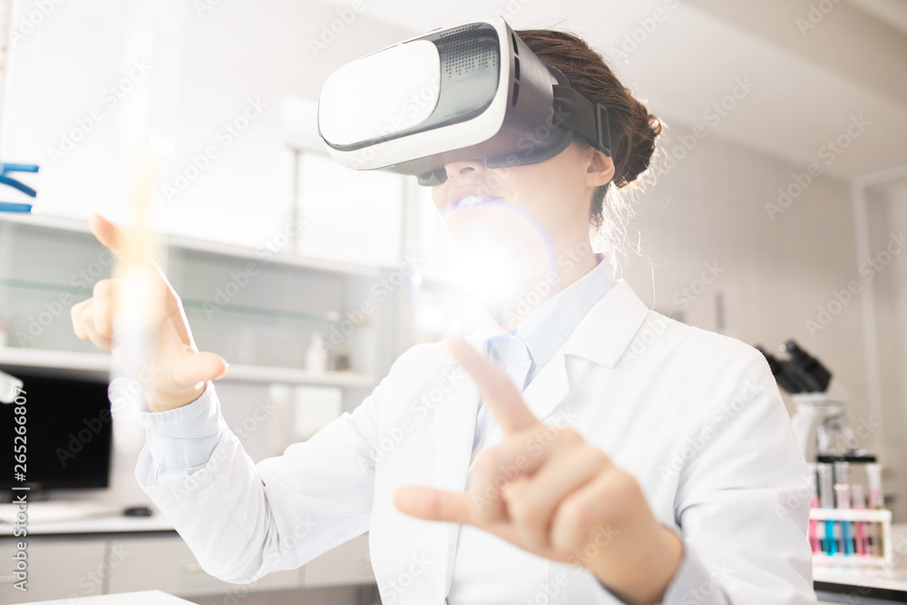 Naklejka premium Positive curious modern young female scientist in white coat using invisible monitor while learning about genes with VR simulator