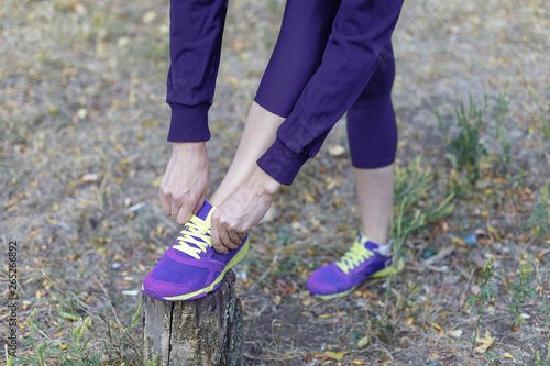 Female legs. Woman in dark violet sportswear ties bright lilac sneakers with lime shoelaces, preparing for a jog, run or other fitness. Morning workout on outdoors nature background. Copy space.
